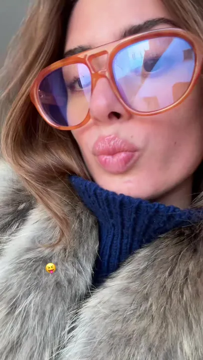 Vinaya in oversized orange sunglasses and a fur collar blowing a kiss